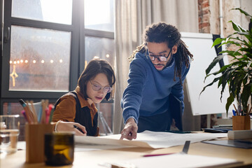 Obraz premium Young Caucasian woman with disability sitting at desk working with blueprints while adult Black man standing beside her pointing at paper, both collaborating on architectural project in modern office