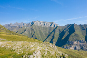Fototapeta premium Bright morning sunlight illuminates a rugged stone escarpment and sweeping green ridges beneath a clear blue sky near Gavarnie Gedre in the Pyrenees. Deep shadows highlight the dramatic contours and