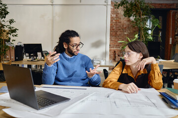 Caucasian young adult woman and Black young adult man discussing architectural blueprints at desk with laptop in modern office setting, both wearing eyeglasses and gesturing while talking
