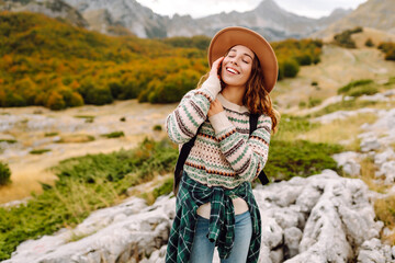 Naklejka premium A young hiker with a backpack and hat on a mountain hiking trail. A smiling woman enjoying the mountain views at sunset. Concepts of weekends, freedom, and adventure.