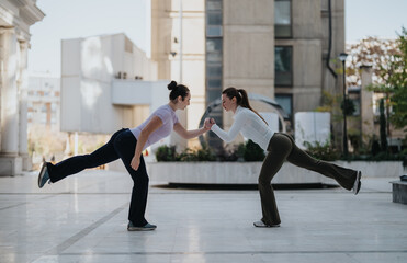 Two women in athletic wear perform a balanced partner workout in an urban plaza. One leans forward...