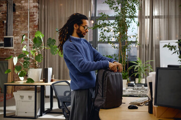Young adult Black man standing in modern office packing backpack on desk, surrounded by computer monitors and large green plants, wearing glasses and having long dreadlocks
