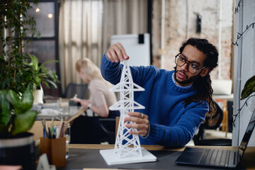 Young adult Black man assembling architectural model of transmission tower at desk in modern office, focusing on precise placement while colleague working in background