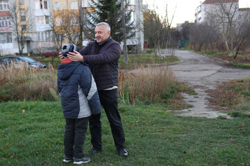 Father adjusting his son's hat during an autumn walk on a grassy area near apartments.