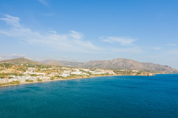 Expansive coastal landscape of Analipsi, Crete, with a sweeping bay, white buildings along the shoreline, and rugged mountains under a clear blue sky. The sunlit scene highlights the vibrant blue