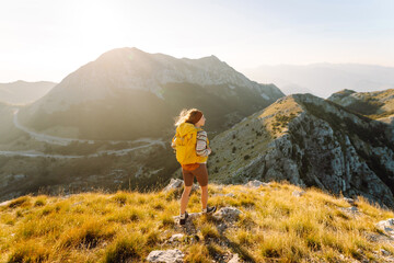 A young woman with hiking backpack stands on mountain trail on sunny day. Female hiker with a yellow backpack enjoys mountain scenery at sunset. Concepts of adventure, freedom. Active lifestyle.