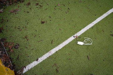 Charging cable lying on a green sports playground ground with a painted white line. Everyday tech object in a casual urban environment, themes of connectivity, technology, and modern lifestyle.