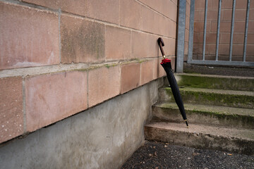 Black umbrella on stone stairs leaning against an industrial wall. Everyday urban scene showing a personal accessory, representing mobility, casual style, and outdoor life.