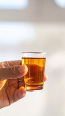 Hand holding a small clear glass filled with amber liquid and golden highlights against a soft blurred background