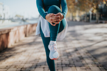 A person in teal active wear lifts a knee in a balanced stretch outdoors. Outdoor workout in a sunny park setting, showcasing fitness and focus. © qunica.com