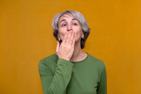 Mature woman expressing surprise or shock by covering her mouth with a hand, isolated on a yellow background
