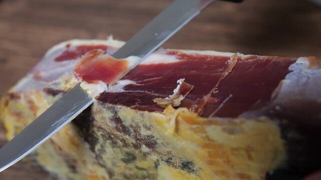 Hand slicing a piece of savory dry cured serrano ham with a sharp knife, carefully carving thin pieces of delicious jamon for a gourmet meal or snack, highlighting traditional spanish cuisine