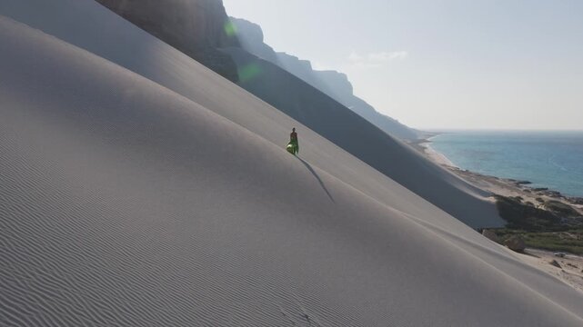 Aerial view of a solitary figure traversing the vast sand dunes of Socotra Island, Yemen, with the tranquil turquoise sea and rugged mountains in the backdrop