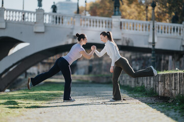 Two women balance on one leg while touching hands in a friendly high-five. They practice outdoor fitness by a river under a decorative bridge.