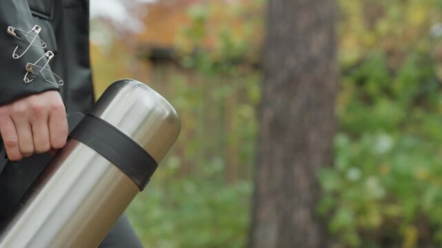 Leather clad white woman carrying thermos, walking through urban forest trail, holding stainless flask by strap, wearing black leather jacket with lace cuffs, tight leather pants and heavy boots,