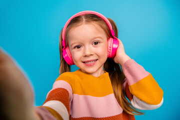 Young girl with pink headphones smiles in colorful striped sweater against blue background portrait...