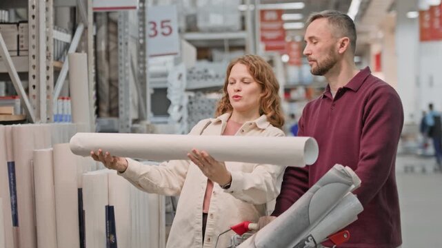 Smiling woman holding roll of wallpaper while discussing it with her husband while standing near showcases