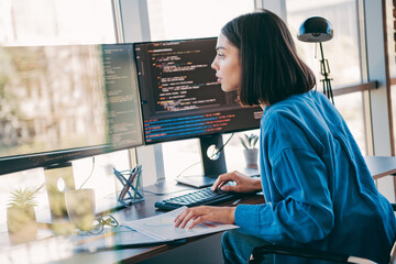 Focused female programmer at a modern desk using dual monitors to work on code in a bright office setting