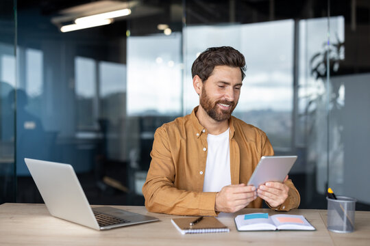Smiling young man with a beard actively engaging with a digital tablet at a desk, surrounded by a laptop, notebooks, and office supplies in a contemporary workspace