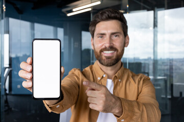 Man holding up a smartphone with a blank white screen for content insertion, pointing a finger towards the display, smiling while standing in a modern office environment