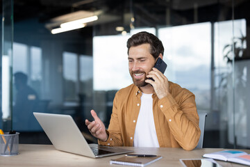 Happy young businessman talking on smartphone while working at laptop in office, smiling and gesturing during a remote call about strategy, productivity and collaboration