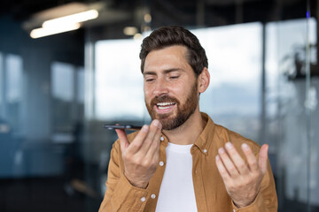 Naklejka premium Man holding smartphone and dictating a voice message, communicating using modern technology in a bright contemporary office setting, showing expression and interaction