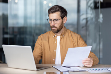 Fototapeta premium Man with glasses concentrating on his modern laptop in a professional workspace, holding and examining financial papers, symbolizing dedication, analysis, and productivity in a corporate environment