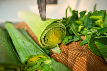 A person is cutting leeks on a wooden board. The green vegetable is fresh and bright, with layers visible