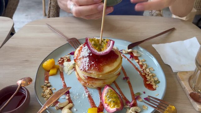 Close-up of a stack of pancakes topped with passion fruit, coconut flakes, and syrup while hands pour sauce. Stylish brunch dish served in a modern caf&eacute; setting