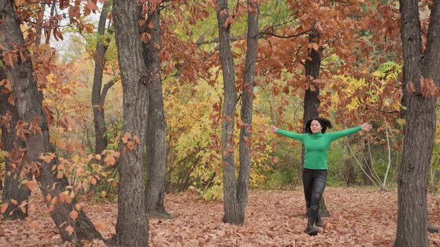 woman in green sweater forest walking between oak trees, leather pants, arms raised in joyful celebration, crunchy orange leaves underfoot, soft overcast light, cinematic slow motion broll capturing