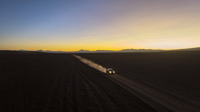 Aerial view of a lone vehicle kicking up dust on a dark road against a vibrant sunset backdrop, horizon mountains, Puna, Jujuy, Argentina.