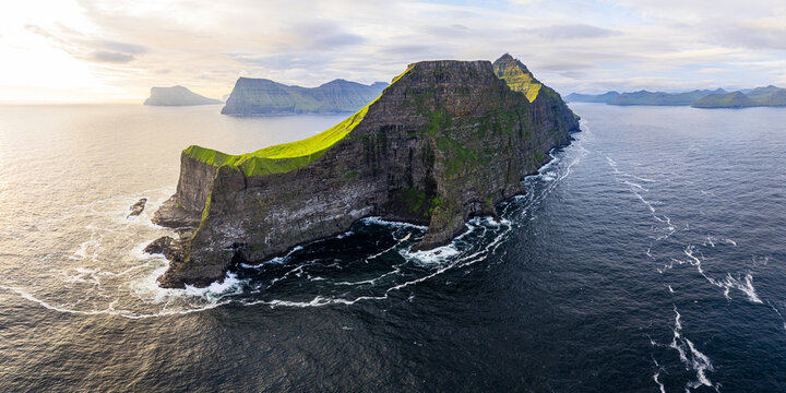 Aerial view of the towering cliffs of Kalsoy (Kallur lighthouse), a dramatic contrast of verdant green slopes against the dark, rugged rock face meeting the churning Atlantic waves, Faroe Islands.