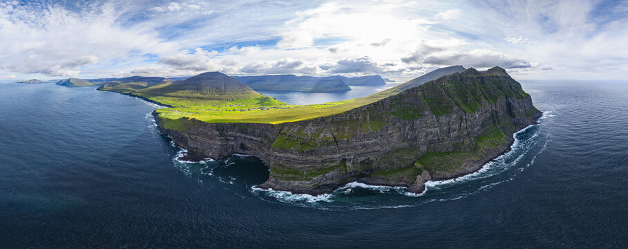 Aerial view of towering cliffs of Vidareidi meet the churning ocean, with verdant slopes and distant islands under a dynamic sky, Vidoy island, Faroe Islands.