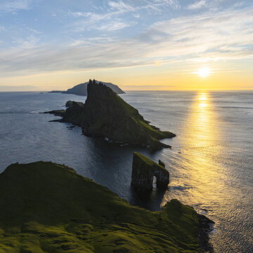 Aerial view of the striking Drangarnir sea stack and Tindholmur islet rise majestically from the dark, swirling waters under a golden sunset, Vagar, Faroe Islands.