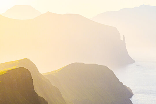 Aerial view of golden light bathing the rugged cliffs of Witches finger and serene waters of the Faroe Islands, creating a dreamy, ethereal landscape, Vagar, Faroe Islands.