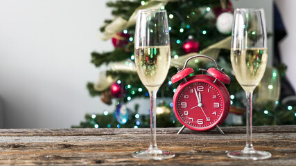 Champagne glasses and red alarm clock in front of a decorated Christmas tree in a cozy indoor setting