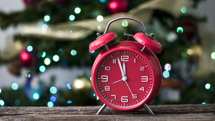 Bright red alarm clock sits on wooden table in front of a decorated Christmas tree with colorful lights in a home setting