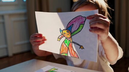 A child proudly shows a colorful drawing to the camera. Captured from a low angle, the video conveys joy and creativity in a warm, sunlit room.