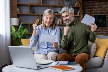 Senior couple receiving good news through mail and online, smiling with excitement and making success gestures while sitting on a sofa in their comfortable living room