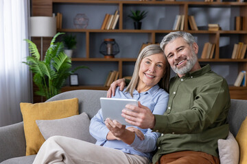 Mature couple smiling at camera while sitting close on a cozy sofa in a modern living room, sharing...