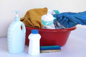 Laundry cleaning concept with gloved hand holding detergent scoop above a red wash basin full of clothes. Detergent bottles, brush, and cleaning supplies arranged on white background.