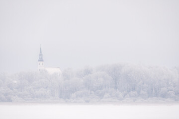 Fototapeta premium Minimalist winter landscape with distant church tower emerging from frosty forest above frozen field, conveying silence, contemplation, spiritual calm and quiet northern atmosphere