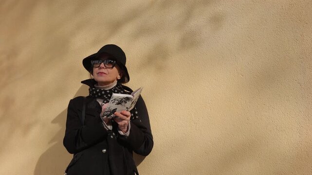Well-dressed mature woman in black coat writes poem in notebook near beige building facade. Inspired senior lady works on romantic story as stands on autumn city street. Creative hobby