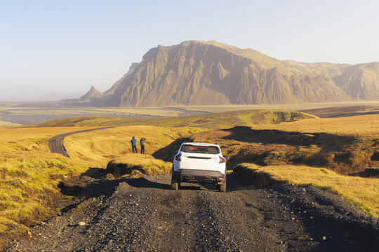 View of a white car traverses a rugged, dark gravel road through golden grasslands toward distant mountains under a clear sky, Myrdalshreppur, Iceland.