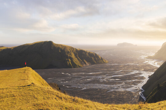 View of golden light kissing the rugged, grassy cliffs overlooking a vast, dark river delta winding its way to the horizon, Sudurland, Myrdalshreppur, Iceland.