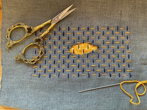 This delicate Sashiko repair transforms a torn hole into an eye-shaped focal point, elegantly stitched with precise running stitches in deep navy and warm golden-yellow thread on a textured fabric