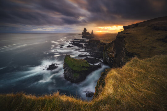 View of dramatic cliffs meet the turbulent sea under a sky ablaze with the fiery hues of sunset, contrasting sharply with the rugged landscape, Arnarstapi, Snaefellsbaer, Iceland.