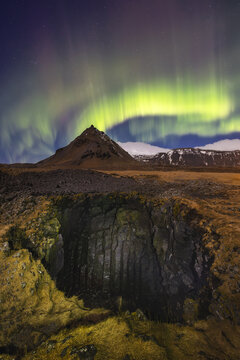 View of the majestic mountain beneath vibrant aurora borealis dances over a rugged landscape with a dark cave, Arnarstapi, Snaefellsbaer, Iceland.