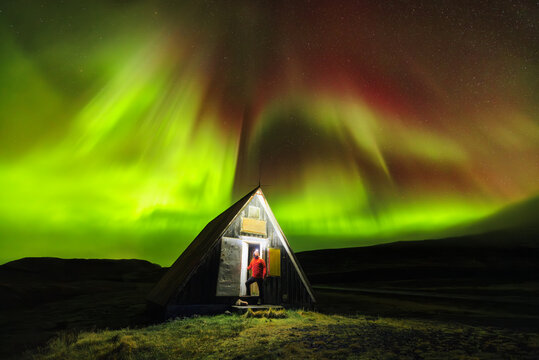 View of a person standing in the doorway of a small triangular cabin under the vibrant, swirling aurora borealis, Arnarstapi, Snaefellsbaer, Iceland.