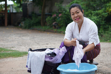 Asian middle-aged woman washing clothes by hand outdoors using a plastic basin. Traditional lifestyle concept showing domestic work, simplicity, and daily routine in a rural environment.